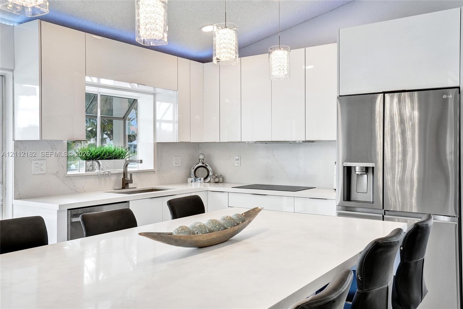 Kitchen featuring white high-gloss cabinetry, stone countertops with matching backsplash, central island, stainless steel refrigerator, and pendant li