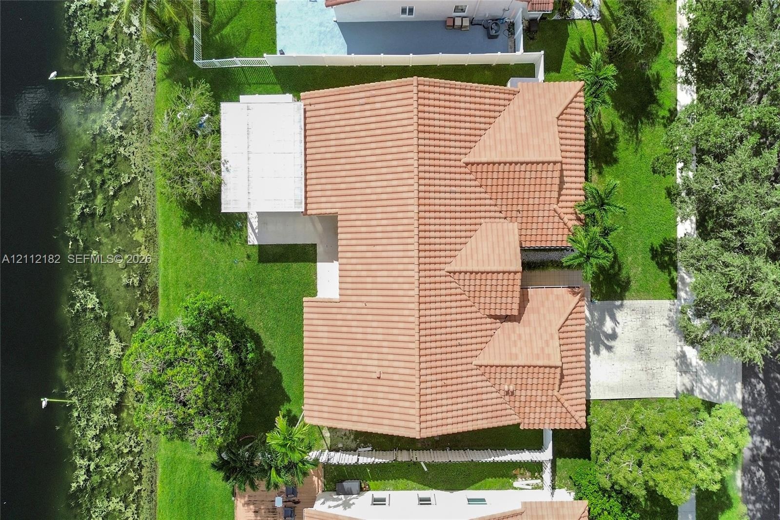 Aerial perspective featuring a tile roof, concrete driveway, attached covered patio, and waterfront lot with mature landscaping