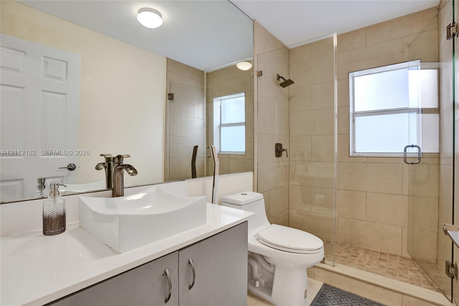 Bathroom featuring a vessel sink with a dark metal faucet, expansive vanity mirror, walk-in shower with floor-to-ceiling neutral tiling, and glass enc
