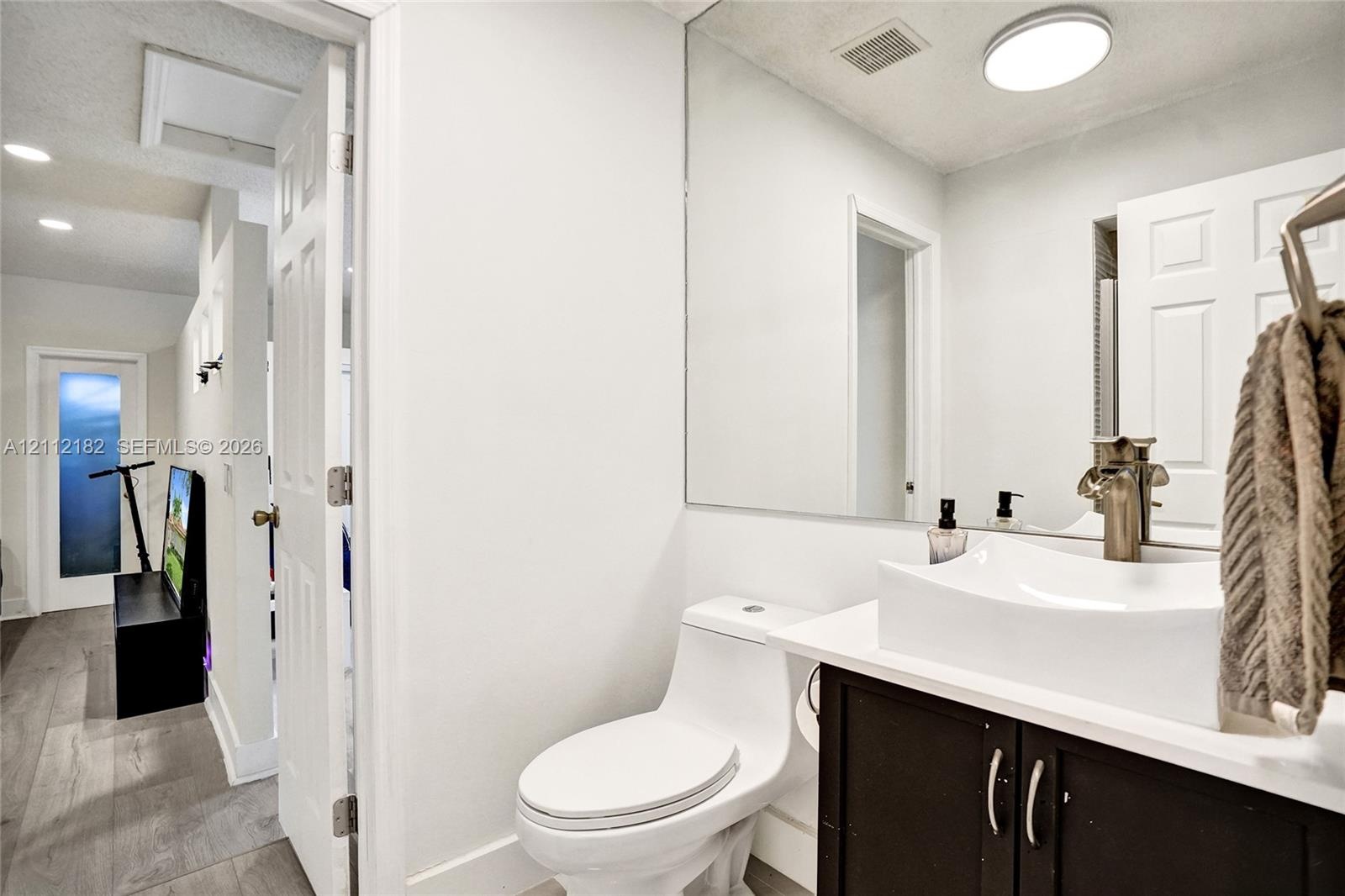 Bathroom vanity featuring a vessel sink, dark cabinetry, and a large-format wall mirror, complemented by wood-finish flooring and a single-piece toile