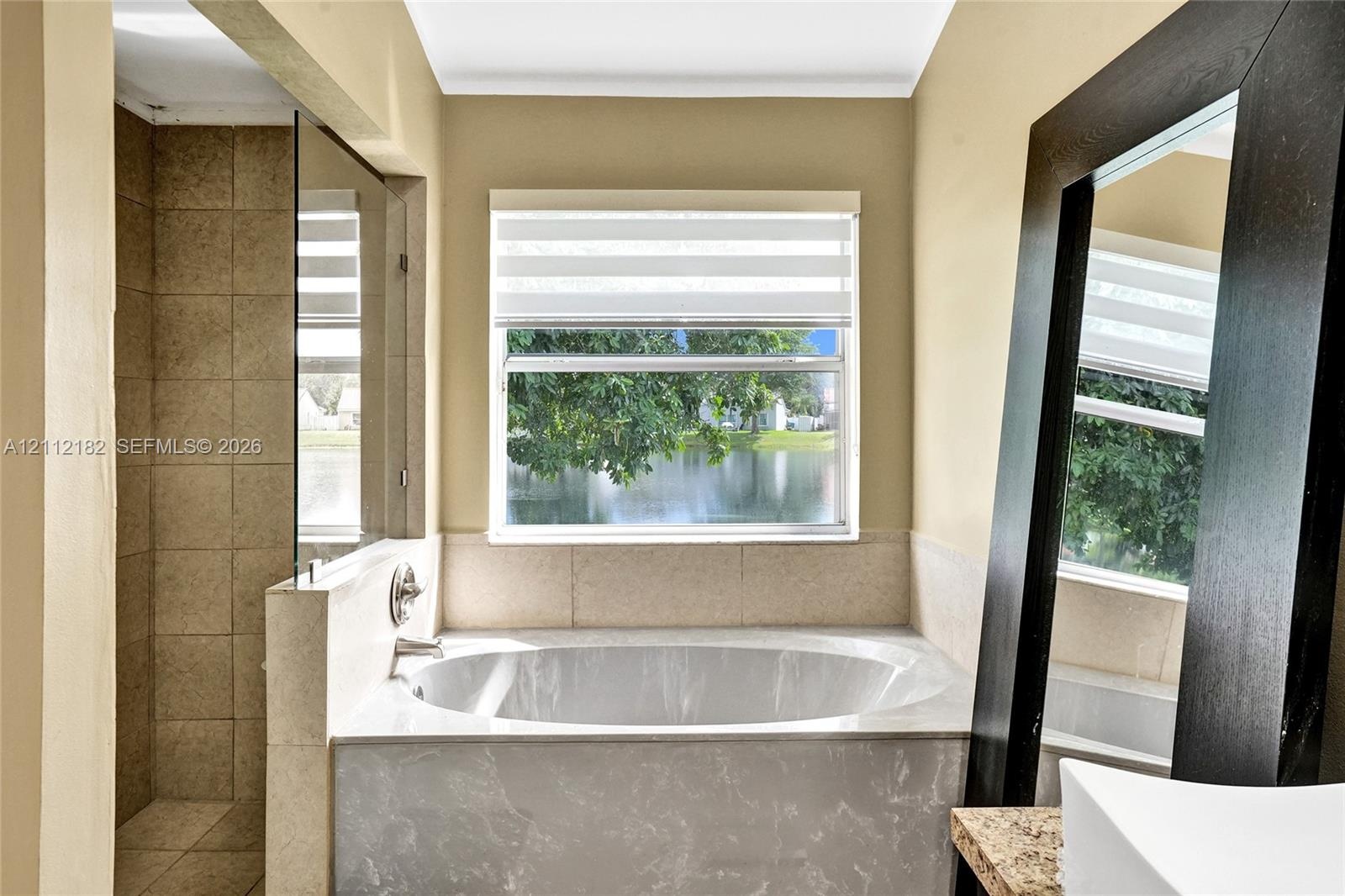 Soaking tub with a stone-finish surround, walk-in shower with floor-to-ceiling tile, and large window with exterior water backdrop
