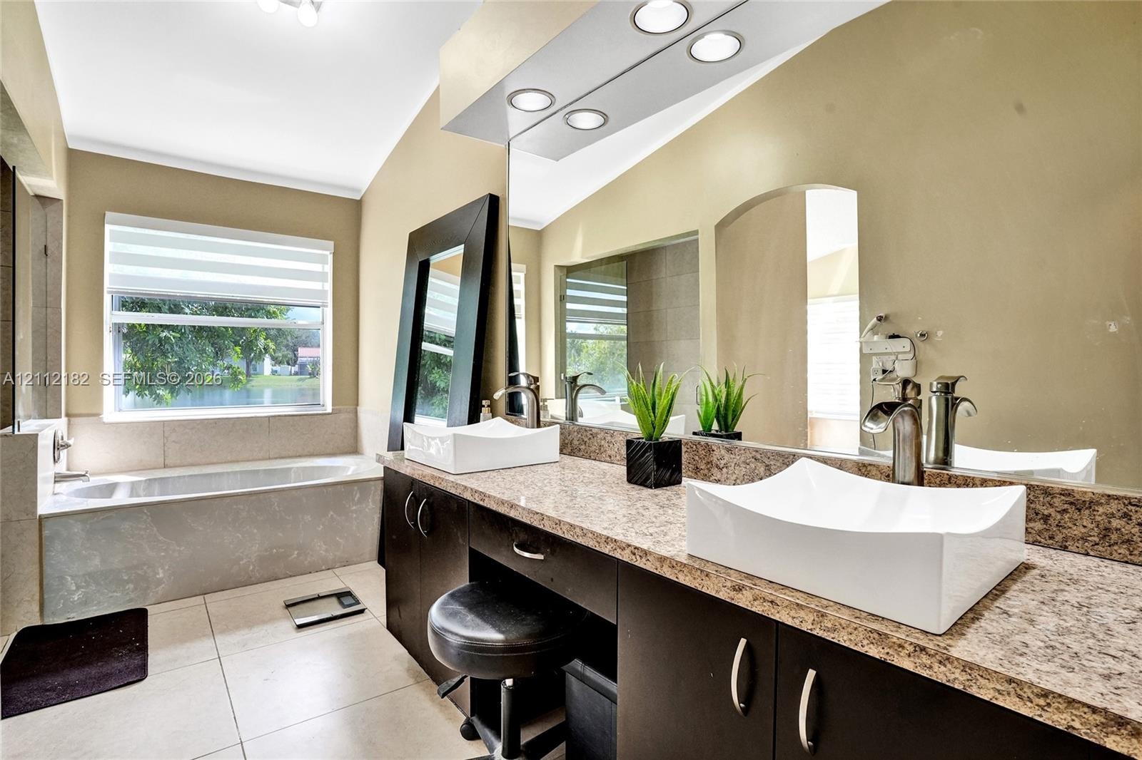 Dual-sink vanity with vessel basins, stone-finish countertop, dark cabinetry, and a deep soaking tub with tile surround