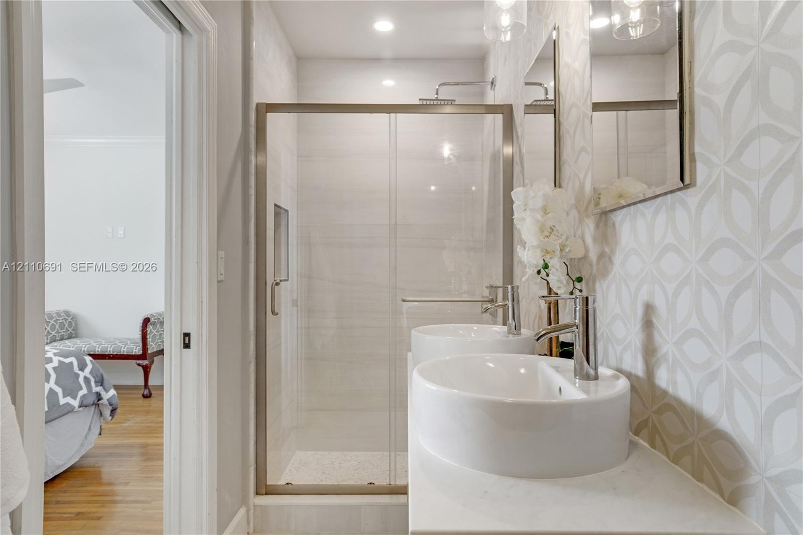 Dual vessel sinks with polished chrome faucets, quartz-surfaced vanity, glass-enclosed shower with recessed shelving, and decorative geometric wall ti