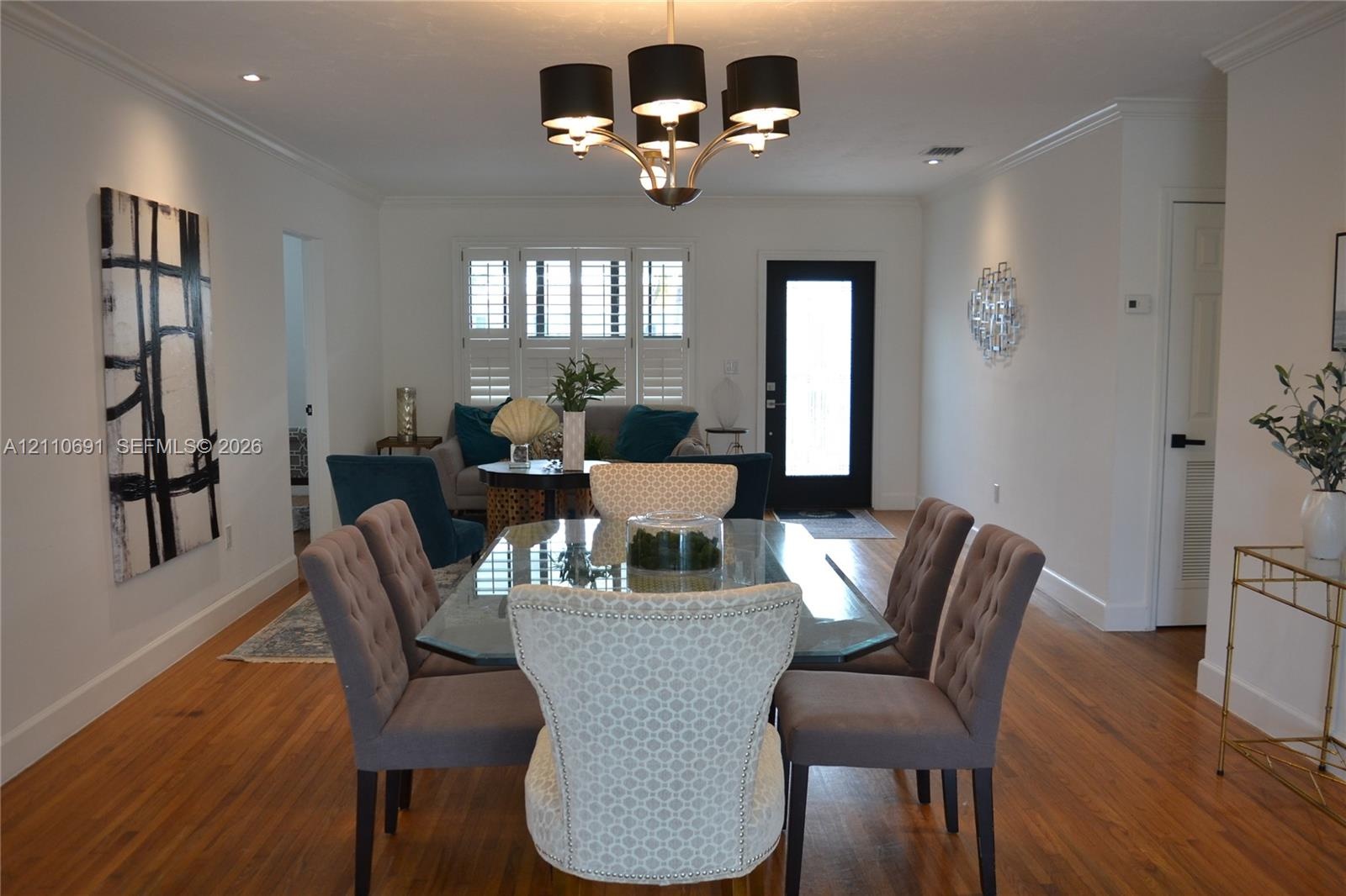 Wood-finish flooring, multi-light chandelier with black shades, white plantation shutters, and a high-contrast black exterior door frame