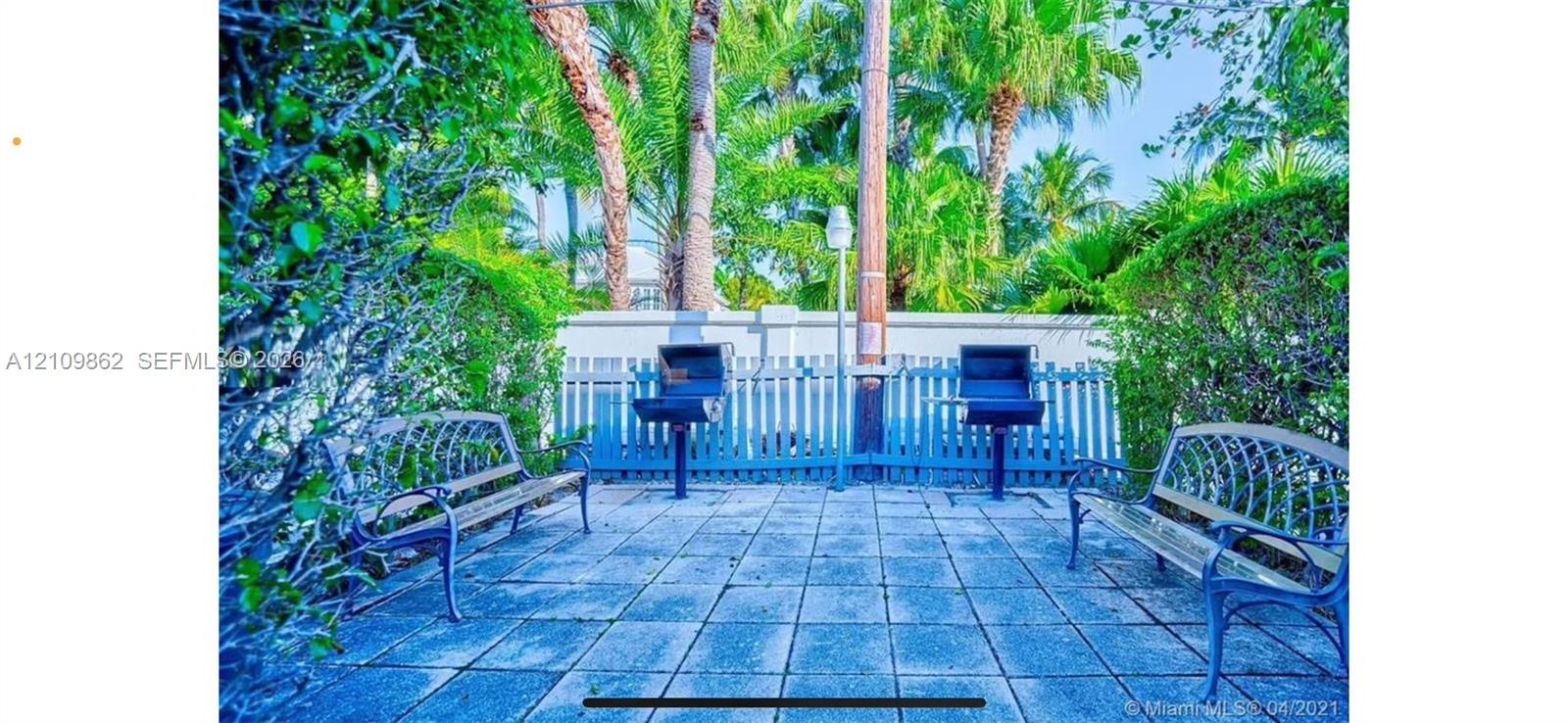 Outdoor patio area featuring square stone pavers, two built-in charcoal grills, and perimeter white picket fencing surrounded by mature trees and gree