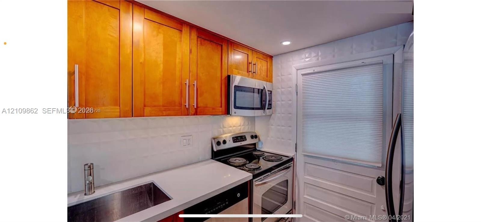 Kitchen featuring wood cabinetry, white countertops, textured white tile backsplash, stainless steel appliances, and a single-basin undermount sink