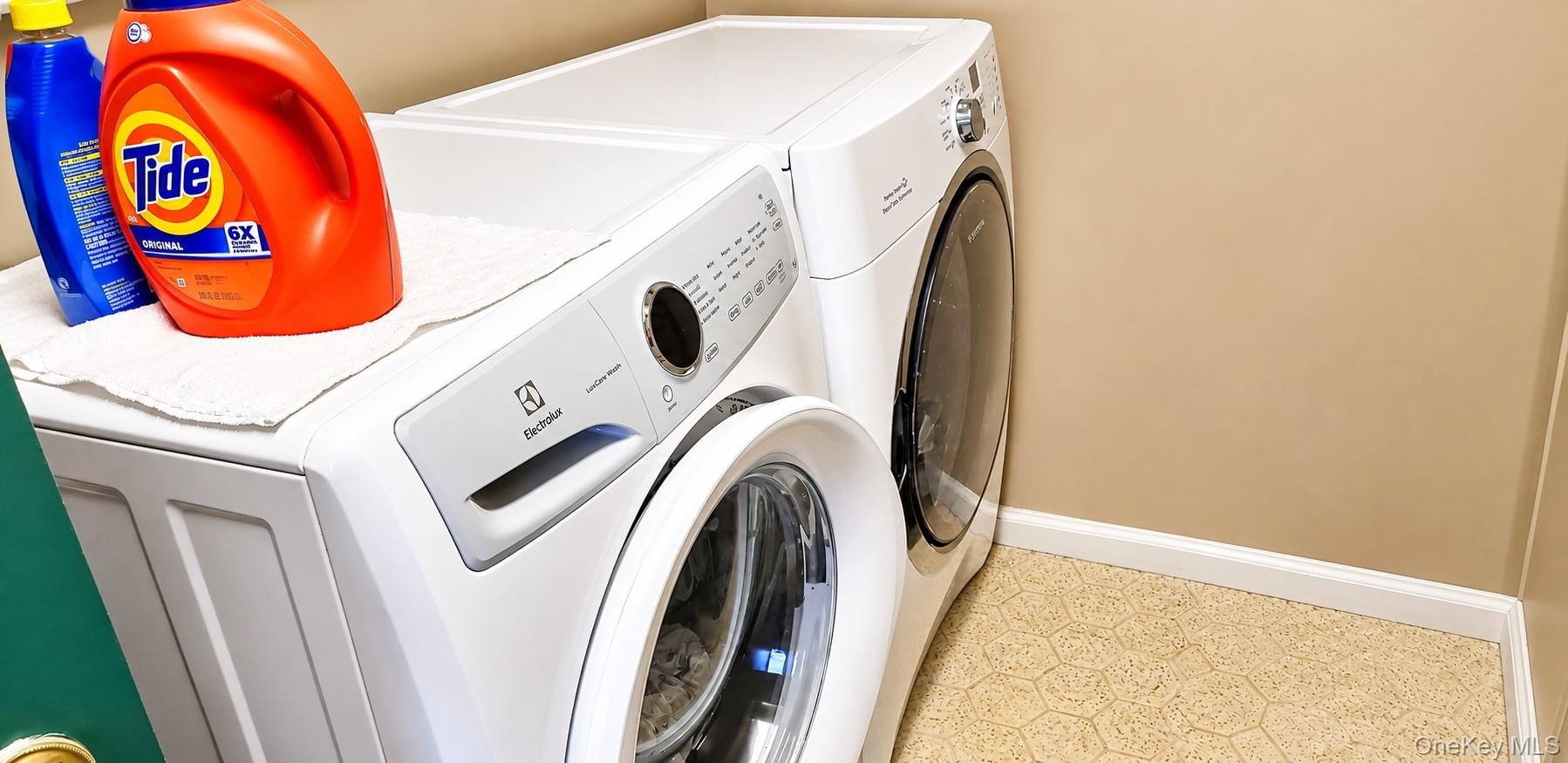 White front-loading laundry appliances featuring digital control panels and integrated glass-front doors, set against a neutral wall finish and hexago
