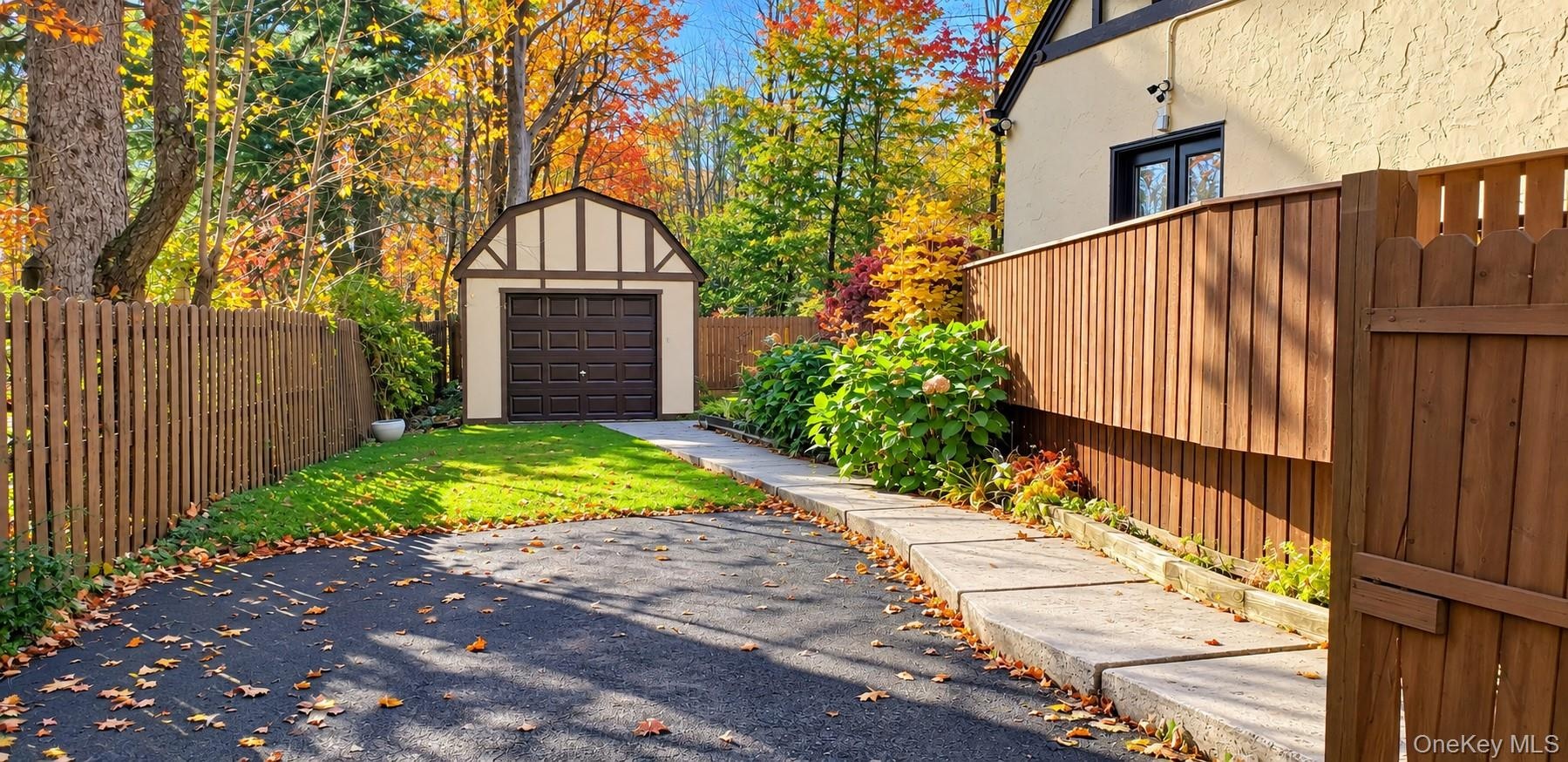 Tudor-style detached garage with stucco and timber detailing, paved driveway, enclosed perimeter fencing, concrete walkway, and landscaped garden beds