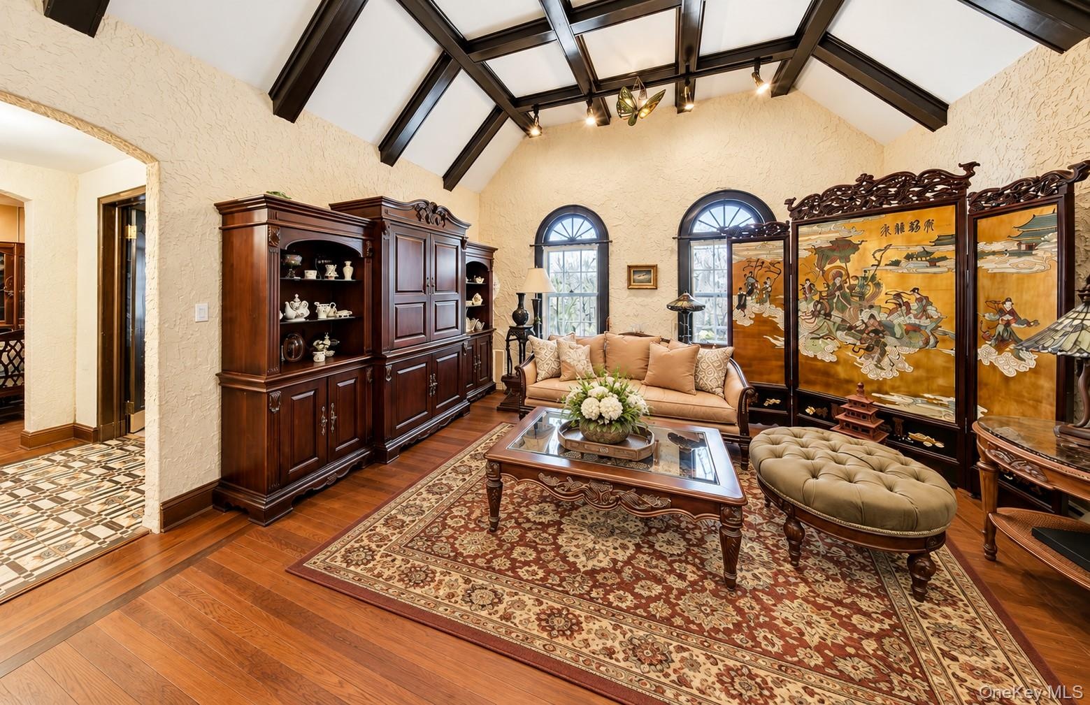 Vaulted ceilings featuring dark exposed beams, built-in wooden cabinetry, arched windows, and wood-finish flooring with inlay borders