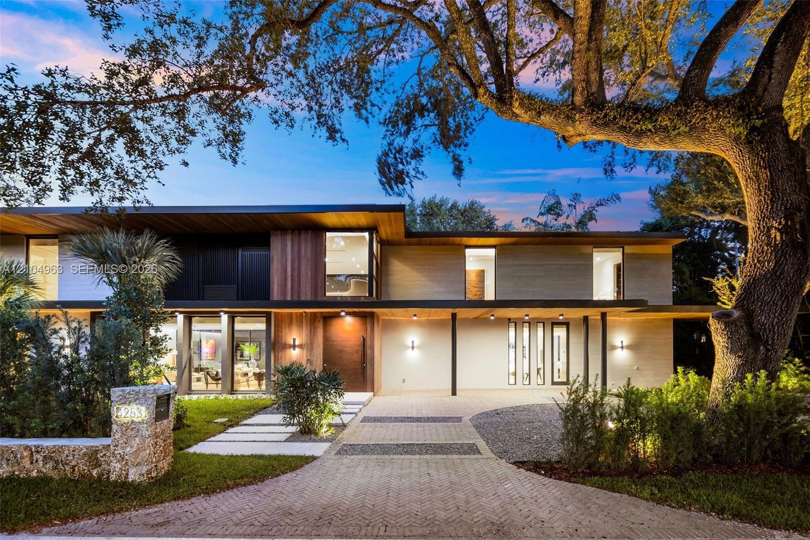 Modern multi-level exterior featuring wood-paneled siding, stone-finished masonry, expansive floor-to-ceiling glass windows, and a paved herringbone d