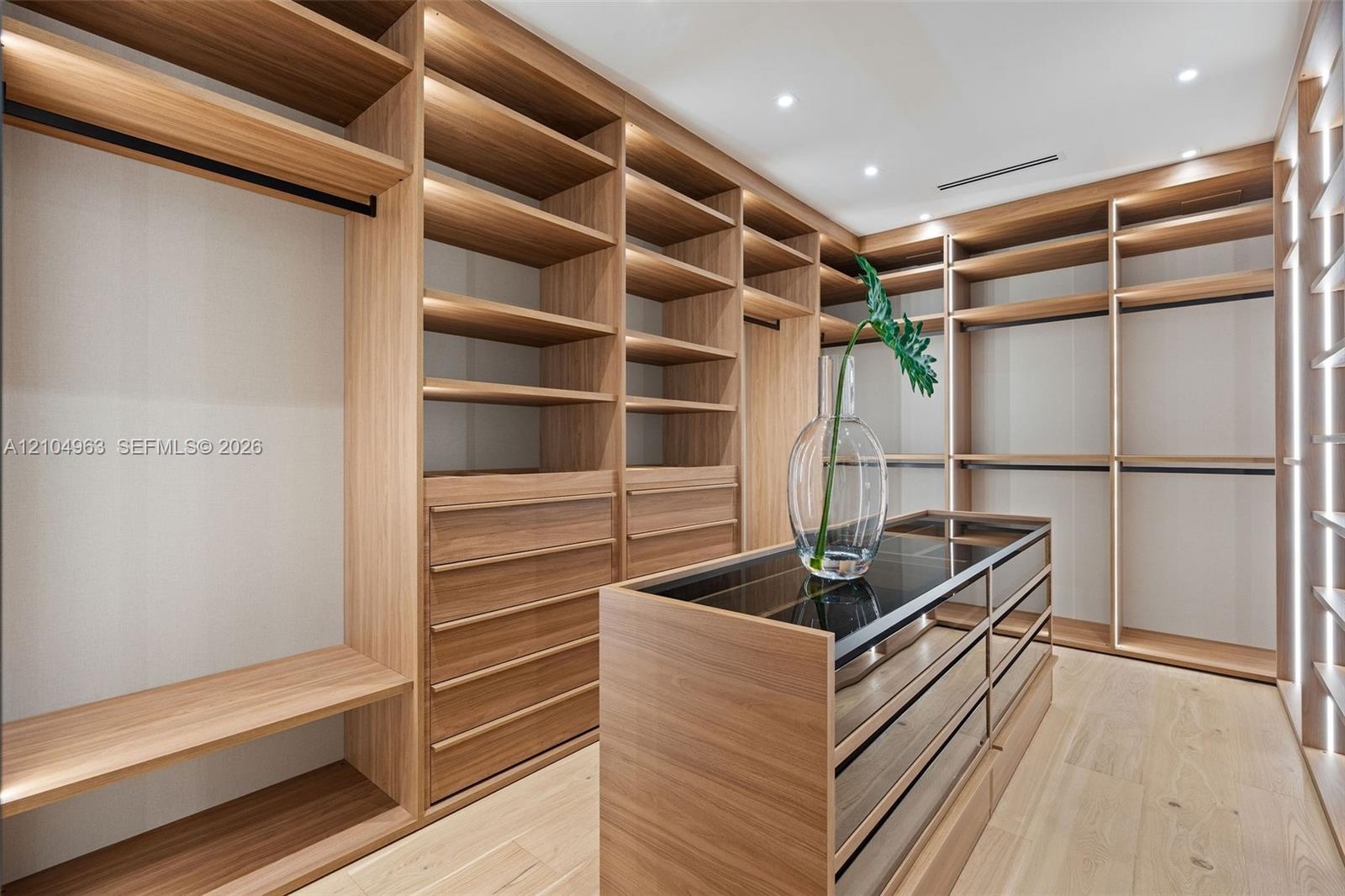 Custom wood-finish cabinetry featuring open shelving and integrated drawers, central island with a glass countertop, and recessed ceiling lighting