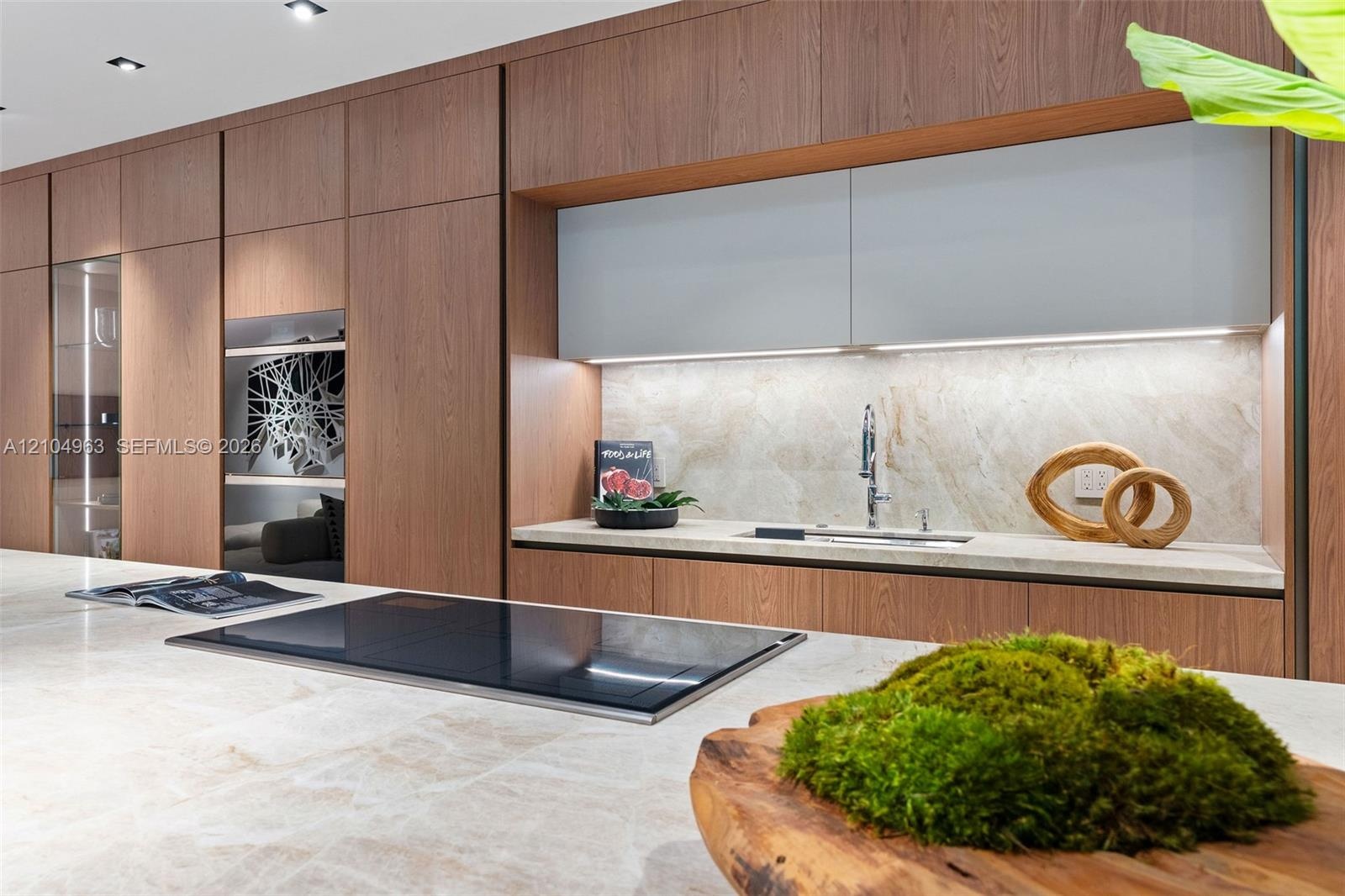Modern kitchen featuring floor-to-ceiling wood-finish cabinetry, a large stone-surface island with an integrated induction cooktop, light-toned stone 