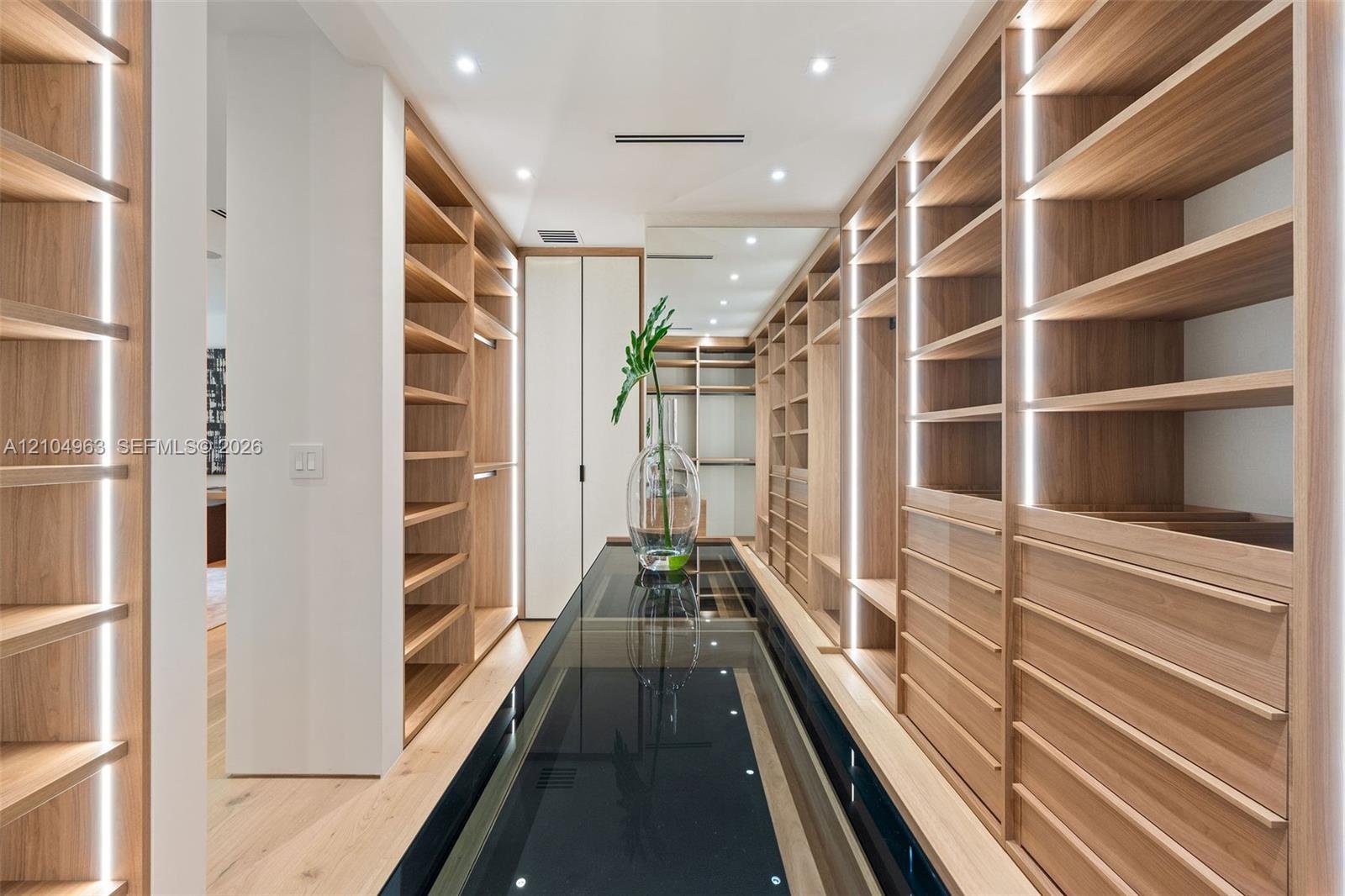 Custom storage cabinetry featuring light wood-finish shelving, integrated vertical LED lighting, a glass-topped center island, and recessed ceiling li