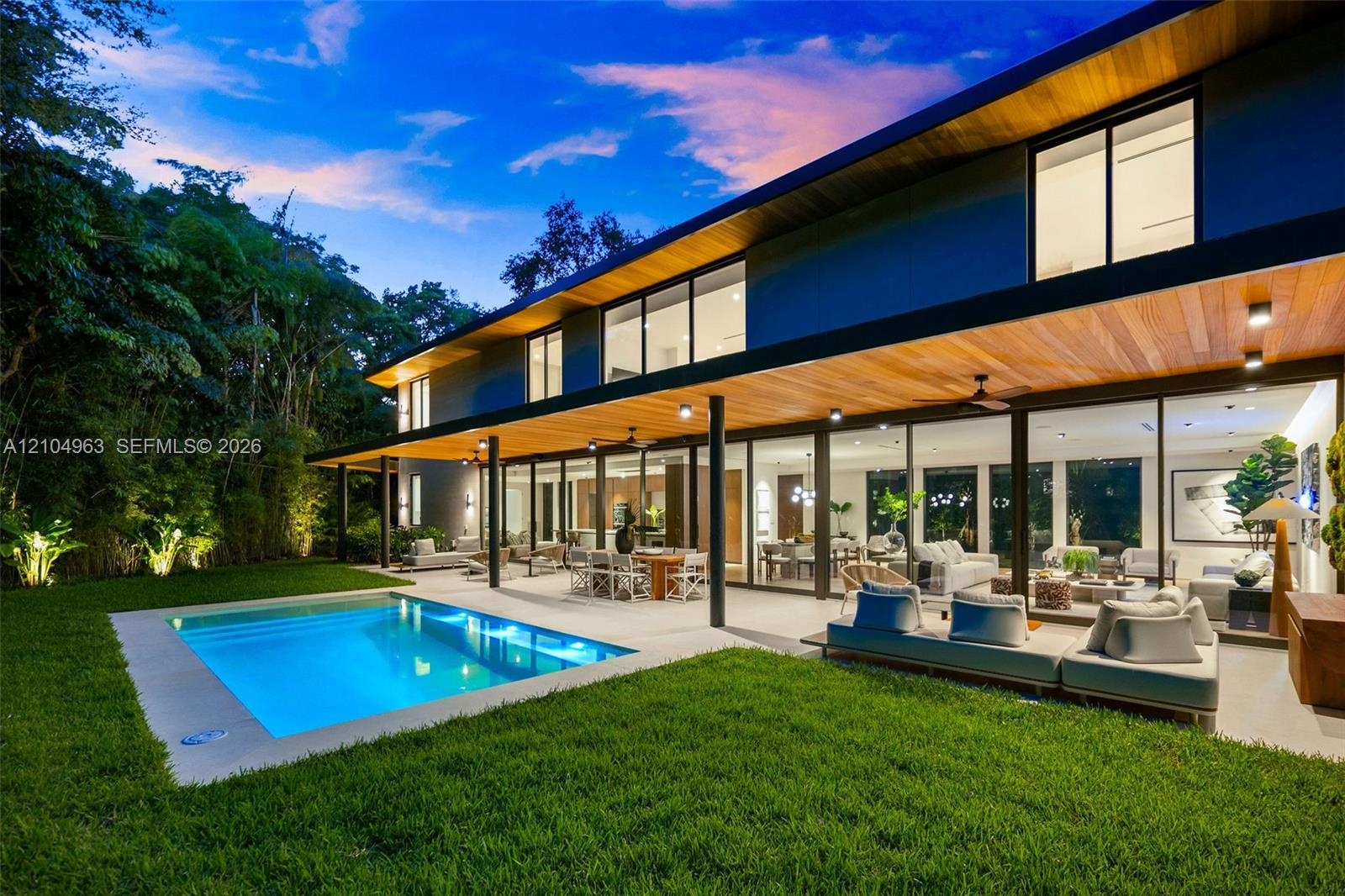 Modern architectural exterior featuring expansive wood-paneled eaves, full-height sliding glass walls, a covered concrete patio, and an in-ground swim