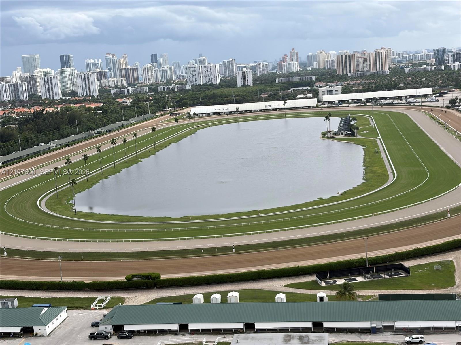 Aerial perspective of an oval racetrack featuring a large central water feature, manicured turf track, dirt perimeter, and established landscaping
