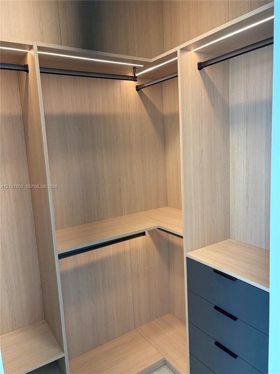 Custom wardrobe system featuring light wood-finish cabinetry, integrated linear LED shelving illumination, matte black hanging rods, and a four-drawer