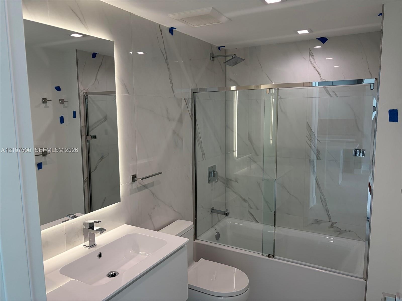 Bathroom featuring floor-to-ceiling large-format tile with decorative veining, vanity with an integrated basin, backlit wall mirror, and a tub-shower 