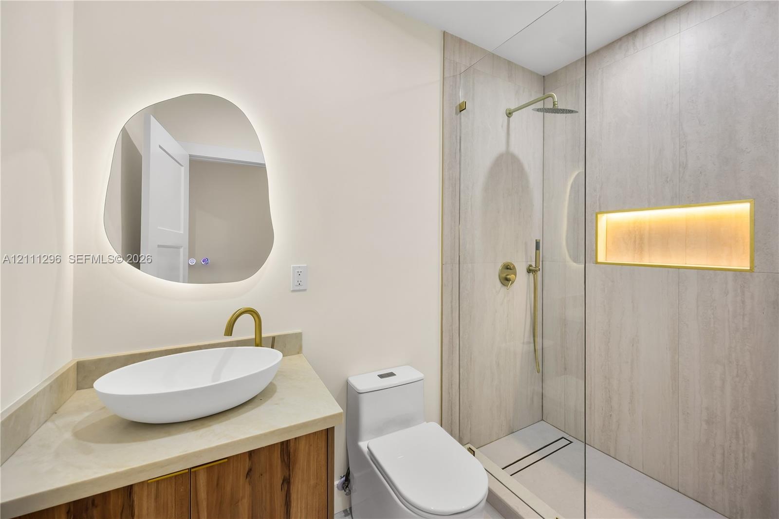 Bathroom featuring an asymmetrical backlit mirror, wood-finish vanity with a vessel sink, brass fixtures, glass-enclosed shower with tiled walls, and 