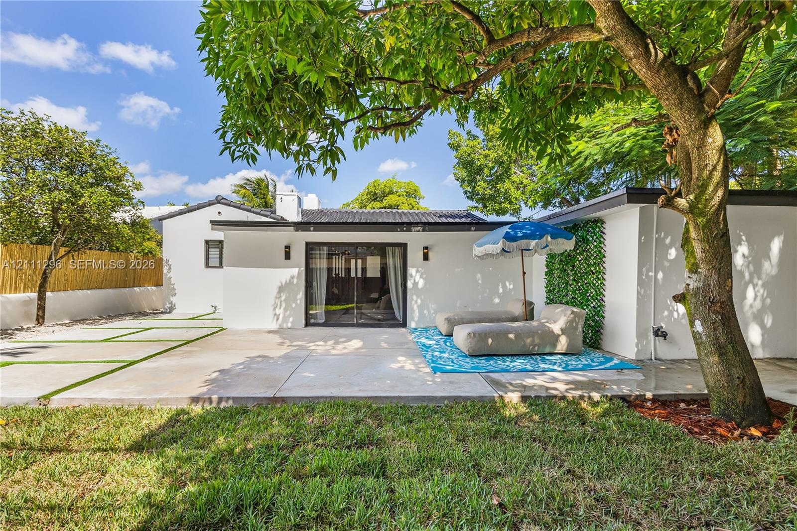 Rear exterior featuring a concrete patio, sliding glass doors, mature trees, a bamboo privacy screen, and exterior wall-mounted lighting