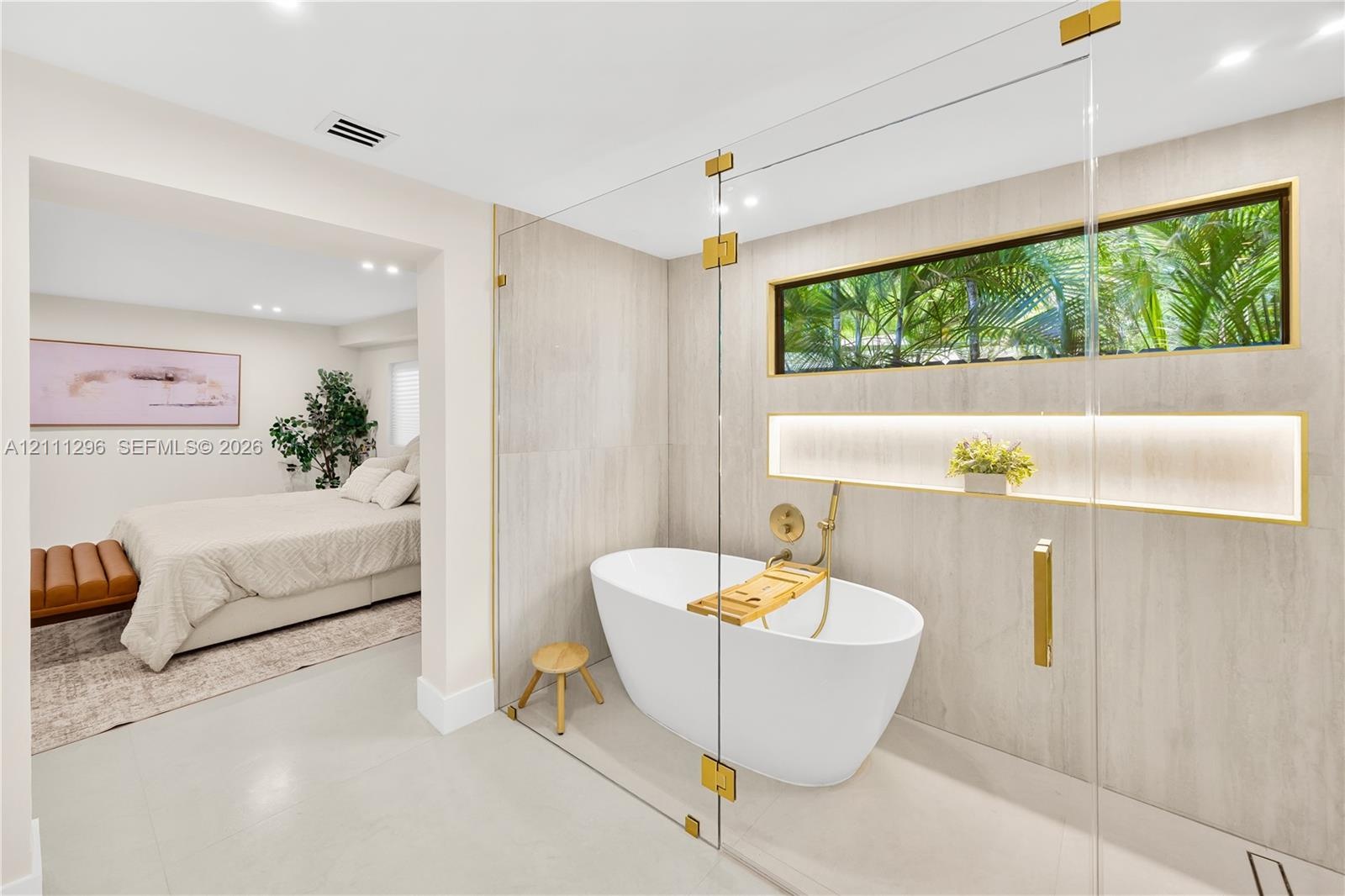 Freestanding white soaking tub enclosed by a frameless glass partition, featuring gold-tone hardware, full-height stone-finish wall tiling, a recessed