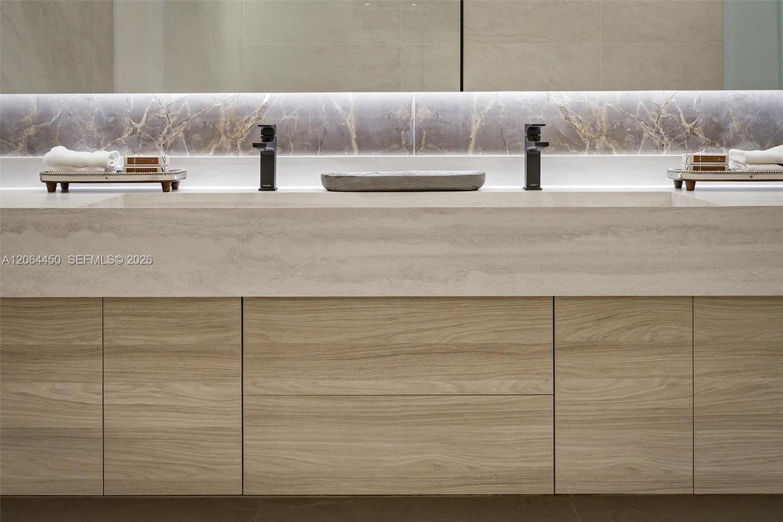 Floating wood-finish double vanity with stone countertop, integrated dual basins, matte black wall-mounted faucets, and polished stone backsplash with