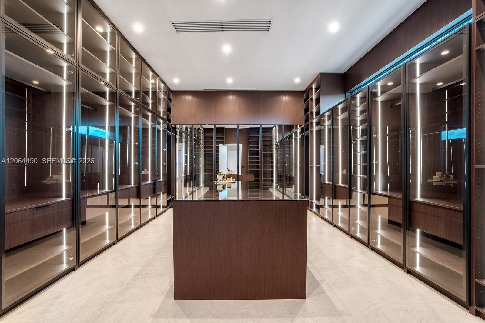Custom dressing area featuring full-height glass-front cabinetry, integrated vertical LED accent lighting, wood-finish millwork, a matching central st