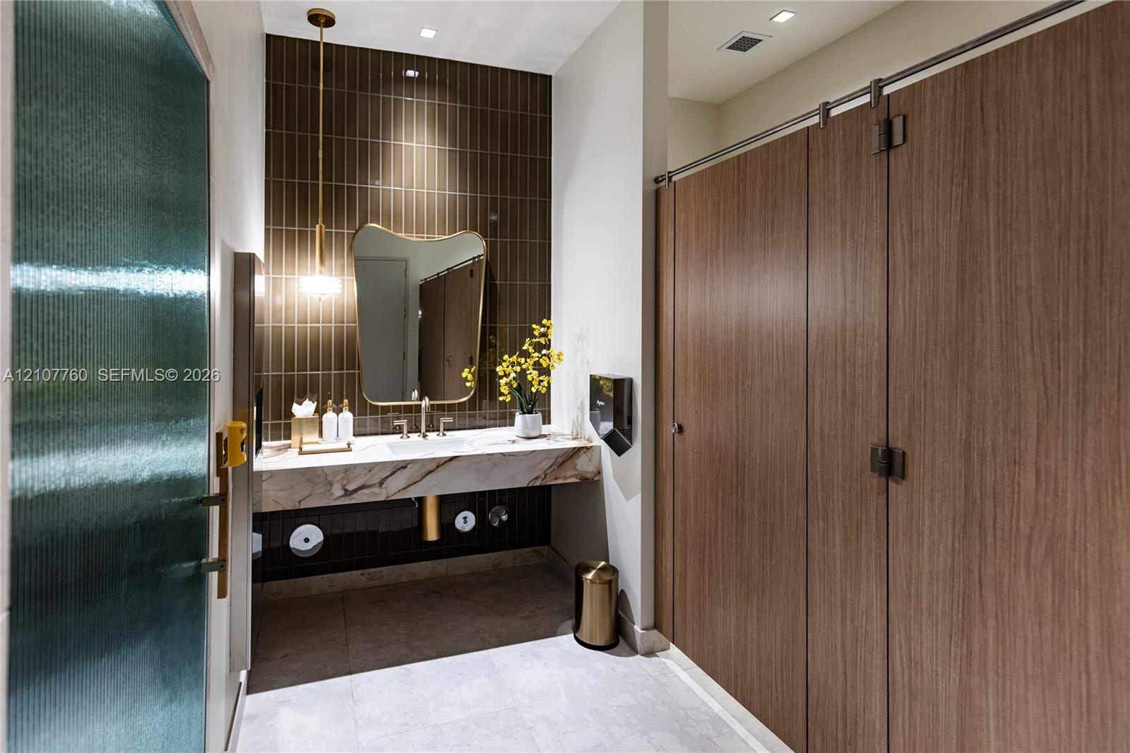 Floating vanity with a stone countertop, vertical tile backsplash, gold-tone pendant lighting, irregular-shaped mirror, and wood-finish partition door