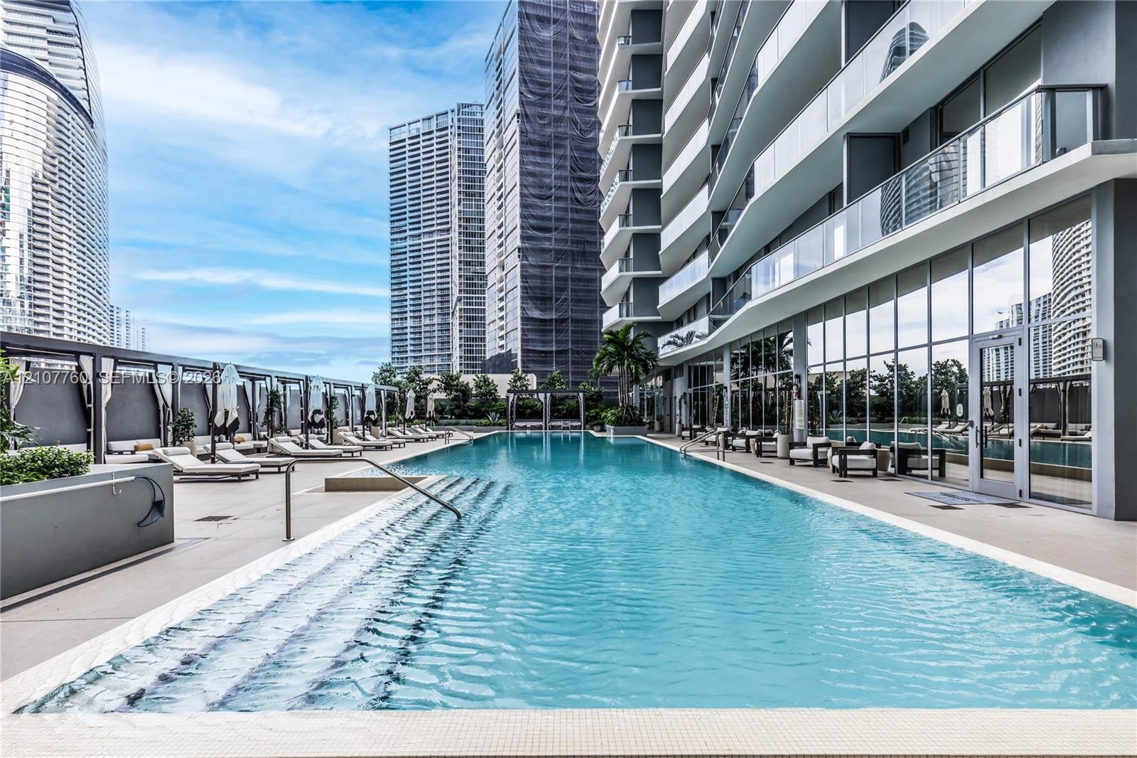 Outdoor lap pool featuring tiled steps, stone-finish decking, glass panel railings, and metal pergolas