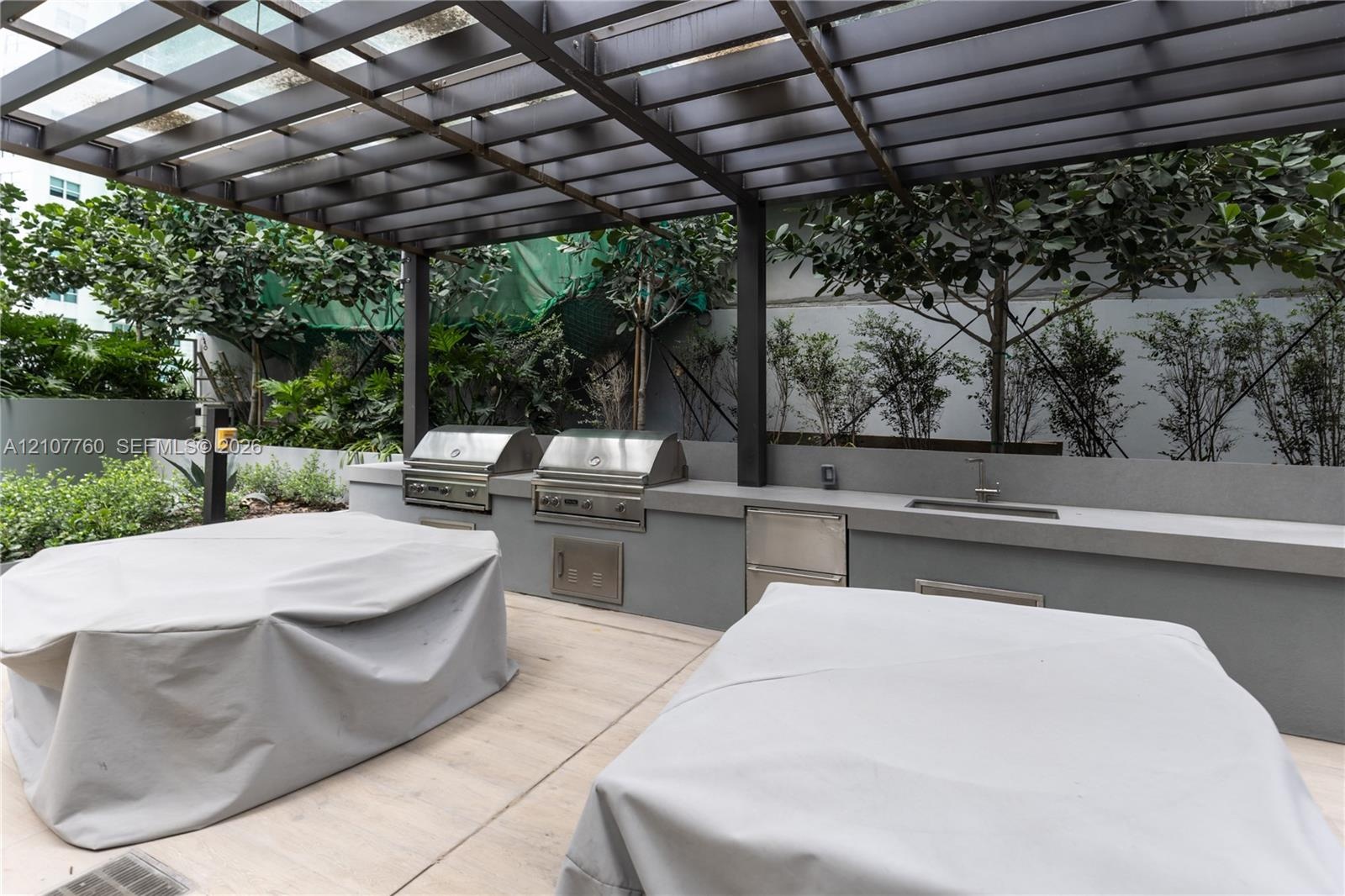 Outdoor kitchen featuring dual stainless steel grills, integrated stainless steel storage, a built-in sink, and a permanent pergola structure set over