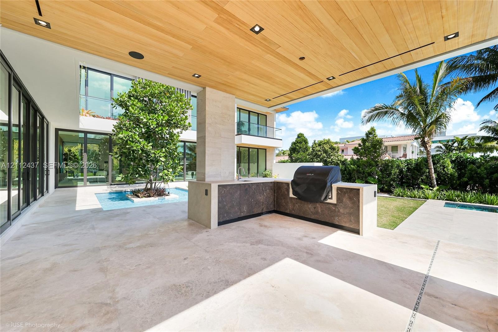 Covered outdoor living space featuring a wood-plank ceiling, natural stone tile flooring, integrated outdoor kitchen with stone countertop, and a cent