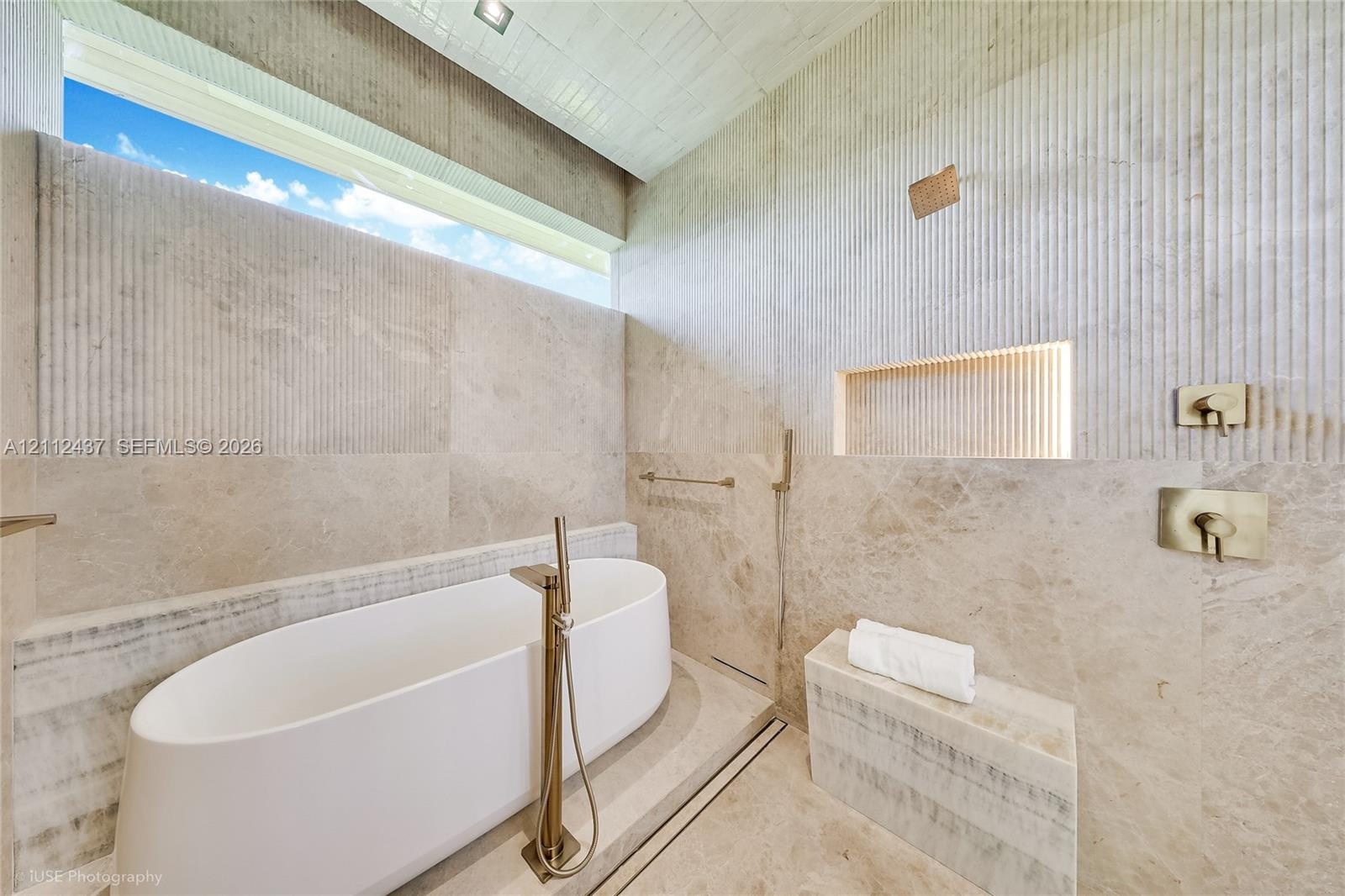 Freestanding white soaking tub with a floor-mounted faucet, floor-to-ceiling stone wall tiling, integrated recessed niche with accent lighting, built-