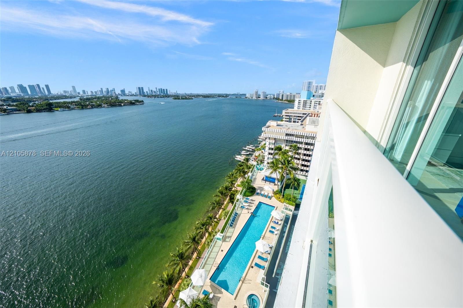 Private balcony with glass railing, overlooking an outdoor swimming pool, hot tub, manicured palm landscaping, and expansive waterfront