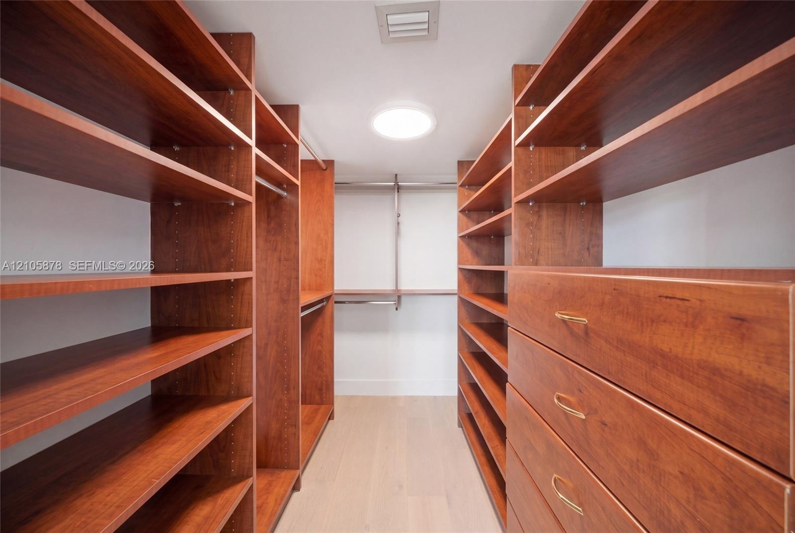 Walk-in storage space featuring extensive custom wood-finish shelving, integrated clothing rods, built-in dresser drawers with metallic pulls, and lig