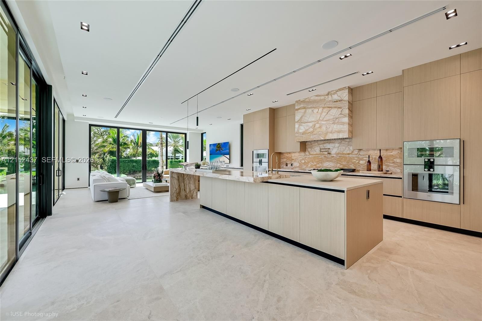 Floor-to-ceiling retractable glass walls, light wood-paneled ceiling with integrated recessed lighting, expansive stone flooring, and custom built-in 