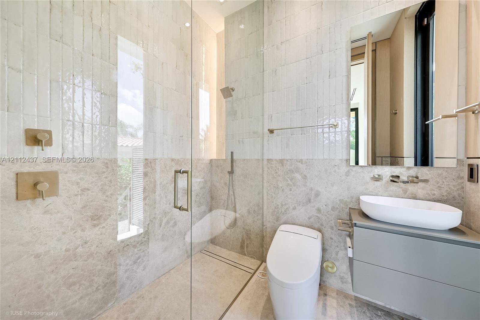 Full-height tiled walk-in shower with a glass enclosure, wall-mounted brass fixtures, stone-finish surround, floating vanity with a vessel sink, and o