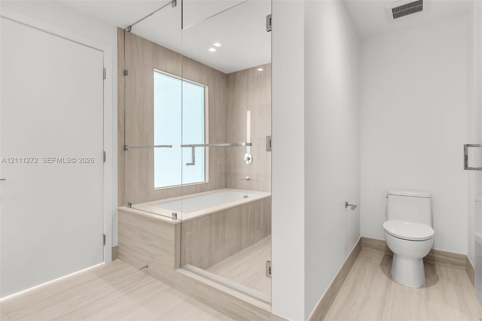 Bathroom featuring a frameless glass-enclosed tub with floor-to-ceiling tile surround, neutral-toned large-format tile flooring, and a white porcelain