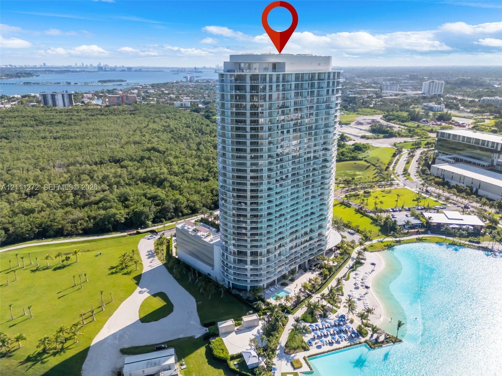 High-rise glass facade building featuring wrap-around balconies, a ground-level swimming pool, and immediate access to a man-made lagoon with a sand s