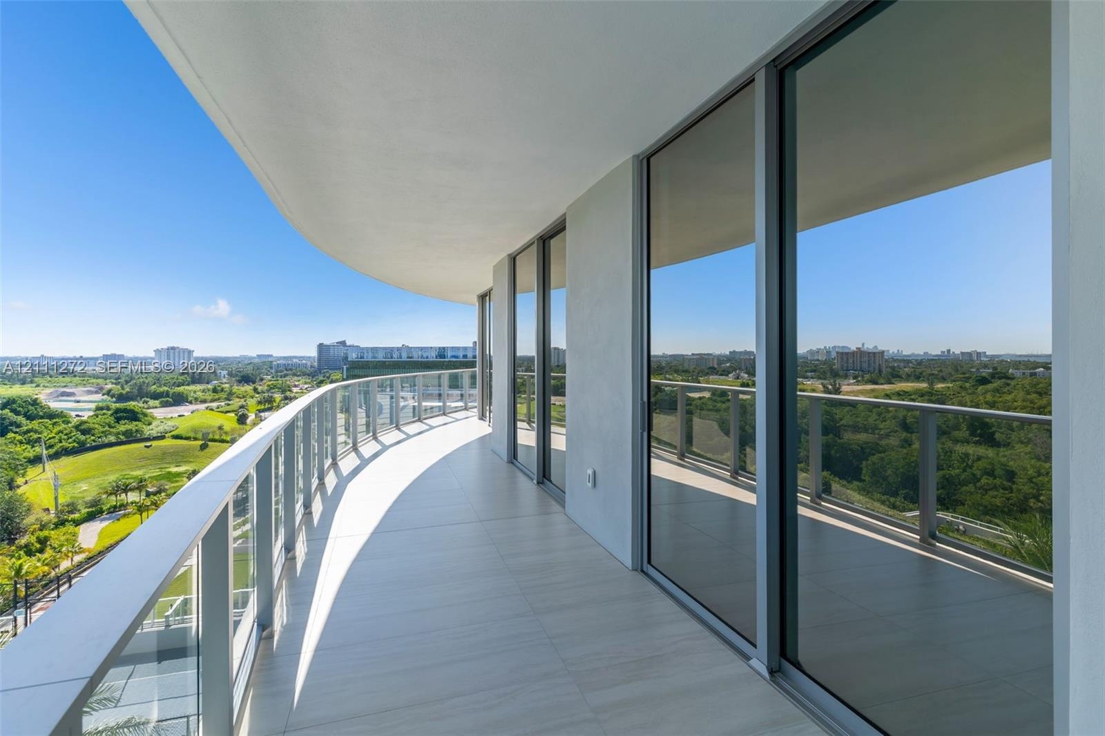 Expansive covered balcony featuring large-format tile flooring, a curved architectural design, and glass panel railing with metal supports, complement