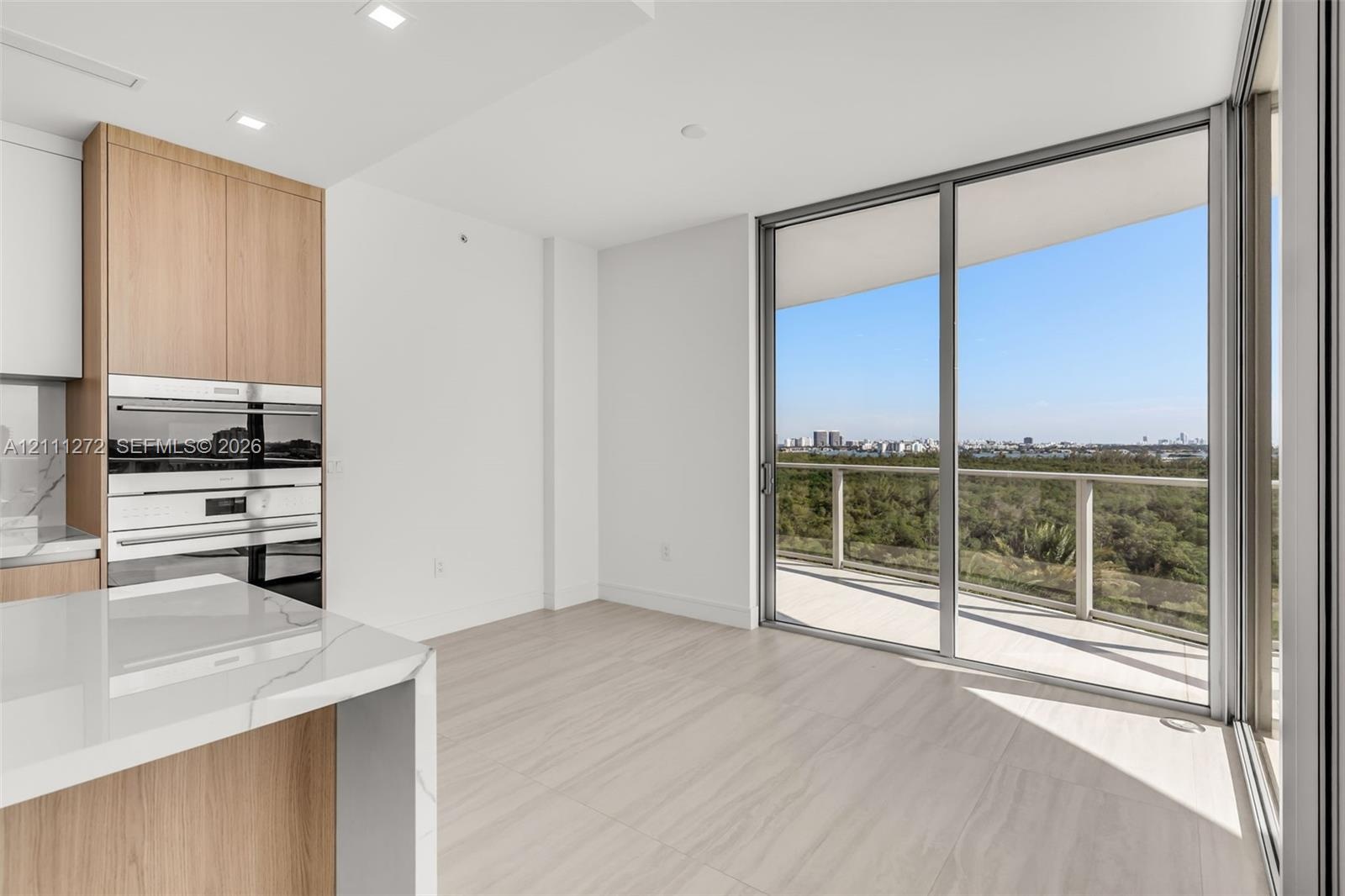 Floor-to-ceiling sliding glass doors opening to a private balcony, wood-finish cabinetry, built-in stainless steel wall ovens, and stone-surfaced isla