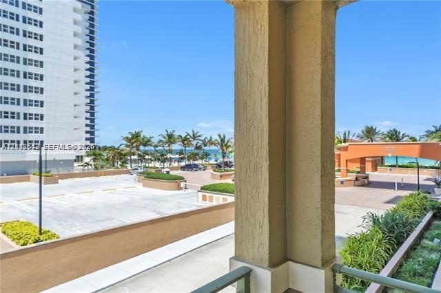 Covered balcony with a textured stucco support pillar, overlooking an exterior surface parking area, landscaped masonry planters, and a coastal landsc