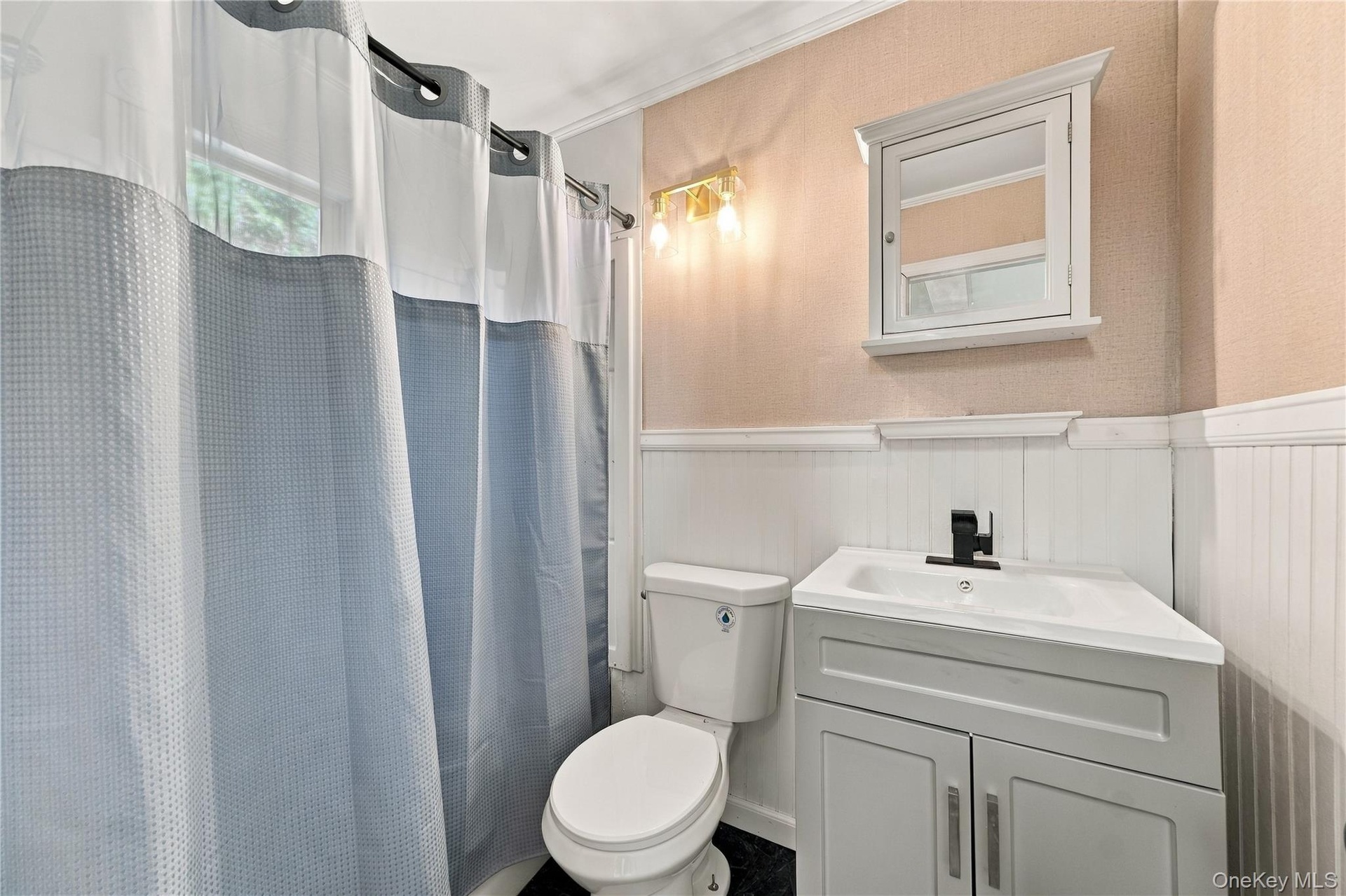 Bathroom featuring a white vanity cabinet with a stone-top integrated sink, matte black faucet, wall-mounted medicine cabinet, and white wainscoting