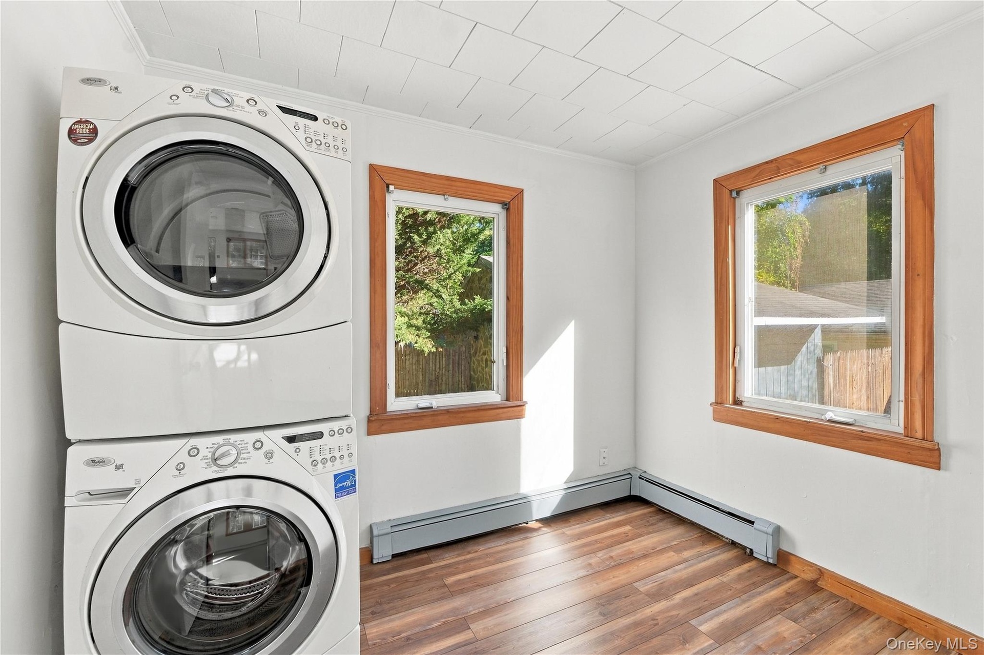 Stacked white laundry appliances, wood-finish flooring, two windows with natural wood trim, freshly painted