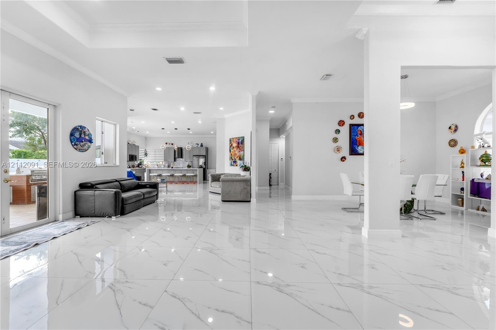 Open-concept main living area featuring large-format polished tile flooring, recessed lighting, tray ceilings, and a glass-paneled door leading to an 
