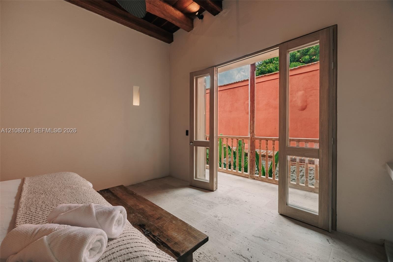 Vaulted ceiling with exposed wood beams, light-toned stone flooring, and full-length glass paneled doors opening to a private exterior balcony with a 