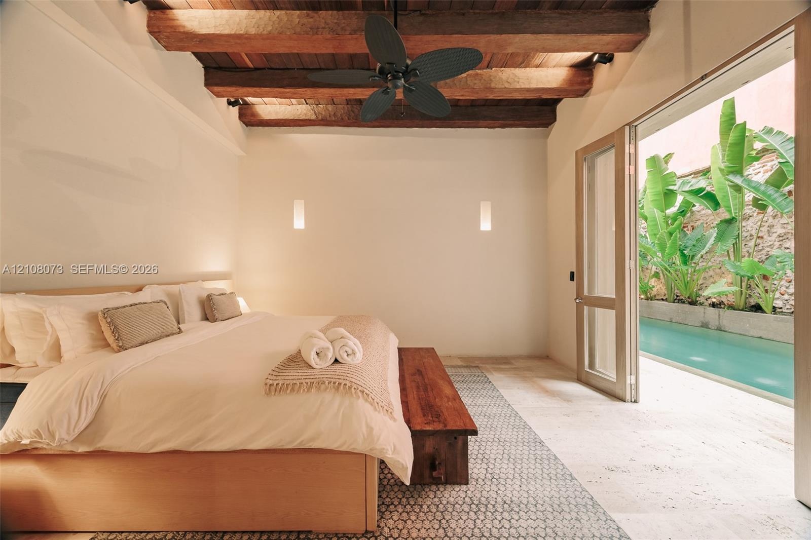 Exposed timber ceiling beams, ceiling fan, wall-mounted light fixtures, stone flooring, and glass door opening to a private pool
