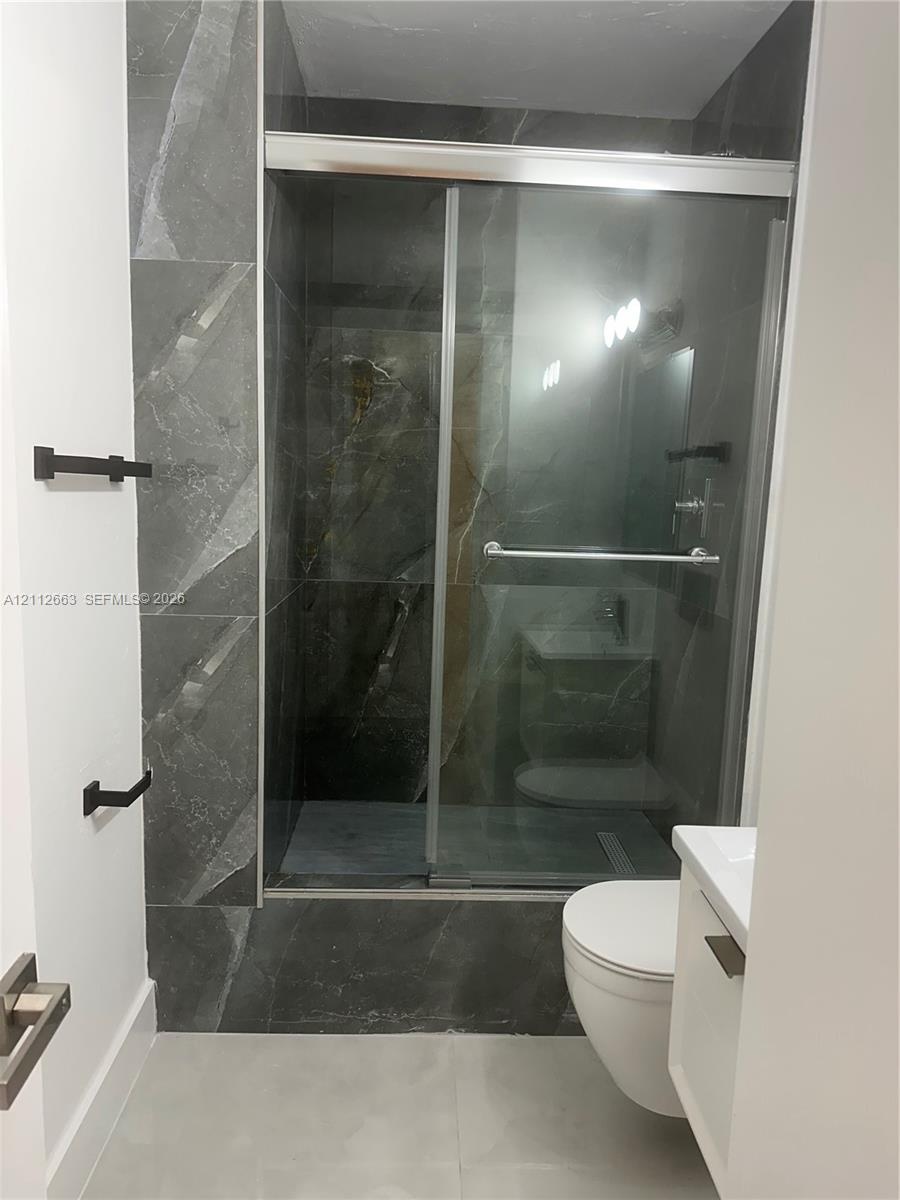 Walk-in shower featuring floor-to-ceiling large-format tile, sliding glass door, wall-mounted matte black hardware, and an elongated toilet