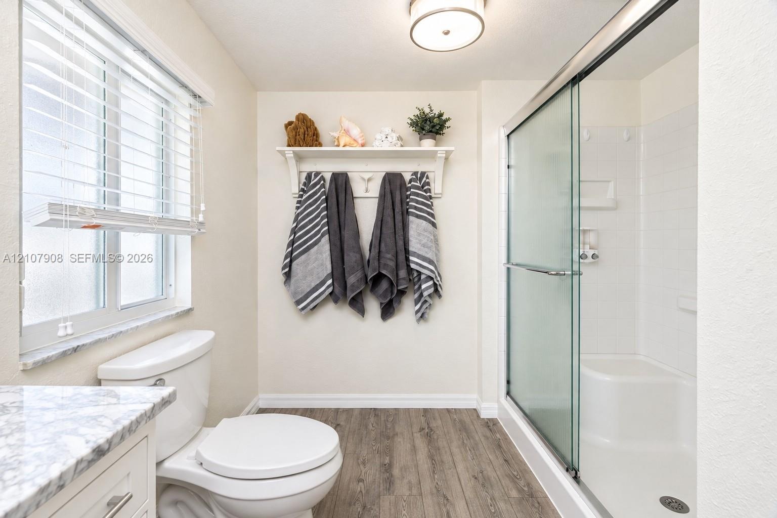Bathroom featuring wood-finish flooring, a walk-in shower with a glass enclosure and built-in bench, stone-patterned vanity countertop, and wall-mount