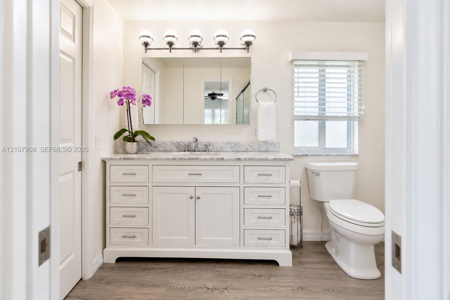 White shaker-style vanity with stone-top surface and undermount sink, multi-drawer cabinetry, chrome five-bulb vanity light fixture, and wood-finish f