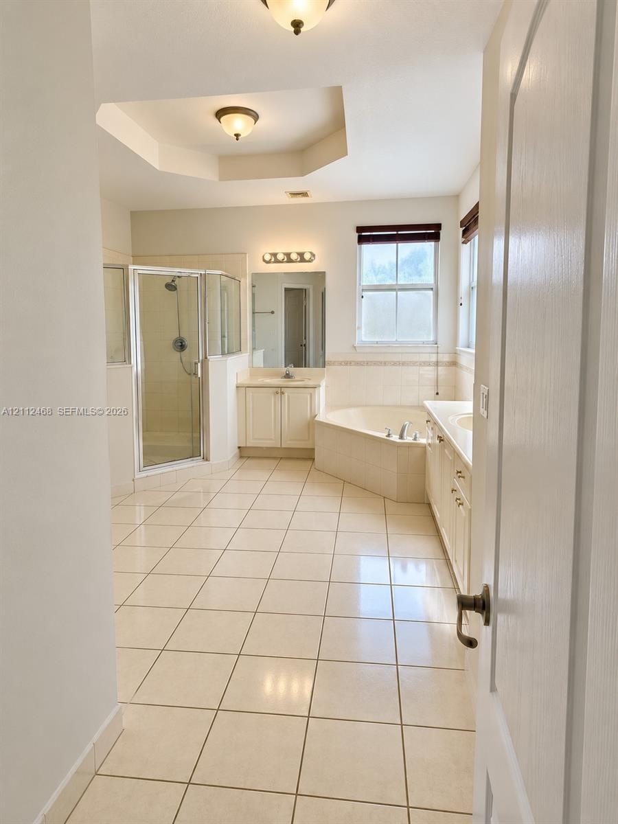 Ceramic tile flooring, corner soaking tub with tile surround, glass-enclosed shower, dual-sink vanity with light-colored cabinetry, and recessed tray 