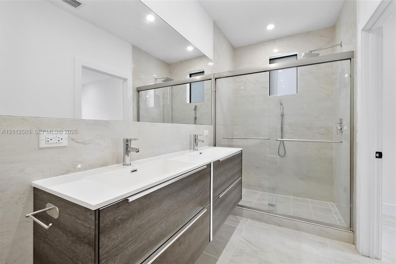 Floating double vanity with a white countertop and dual chrome faucets, complemented by a large wall-to-wall mirror, walk-in shower with sliding glass